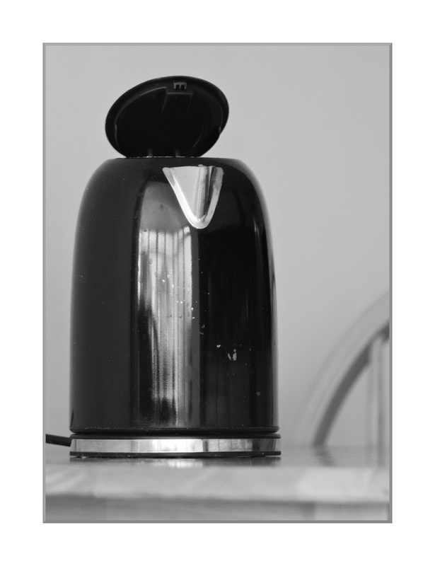 A black enamel. electric kettle, with lid ajar on a table with a part of a chair in the background. Natural window light.